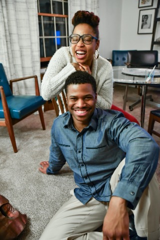 a black couple smiling. The lady is sitting behind the guy on a wooden chair, the guy is sitting on the floor in front of the lady.